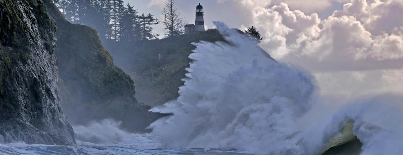 High king tide wave crashes against rocky cliffside with a lighthouse