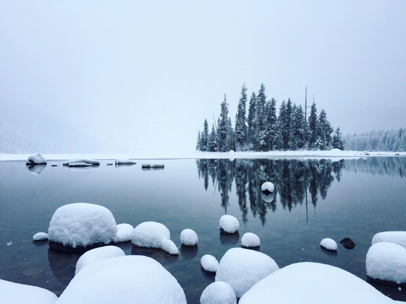 A lake in winter is surroudned by snow. There is a small island with evergreen trees in the distance.
