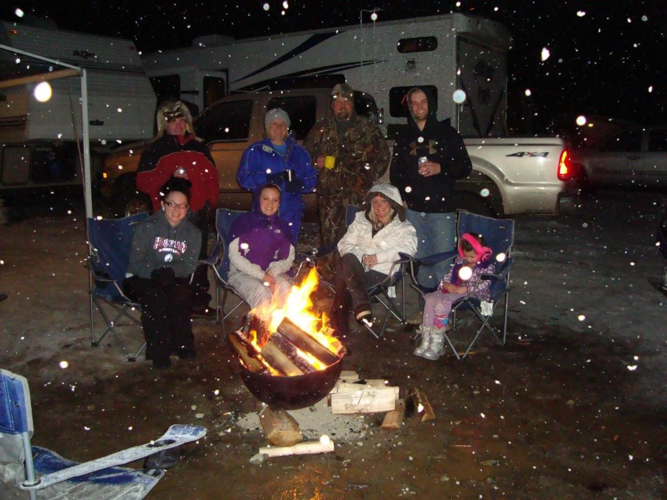 A group of people sit around a campfire while snow falls.