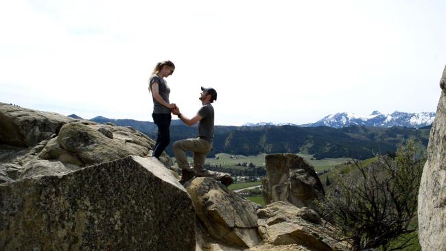 a young man down on his knee on the top of a rock overlooking an orchard proposing to a young woman.