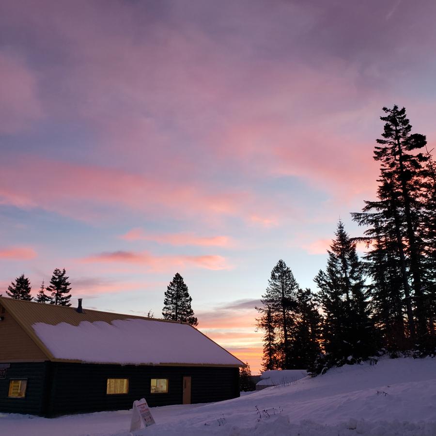 The wooden lodge is covered in snow with a sunset view of trees and the partially clouded sky in the background