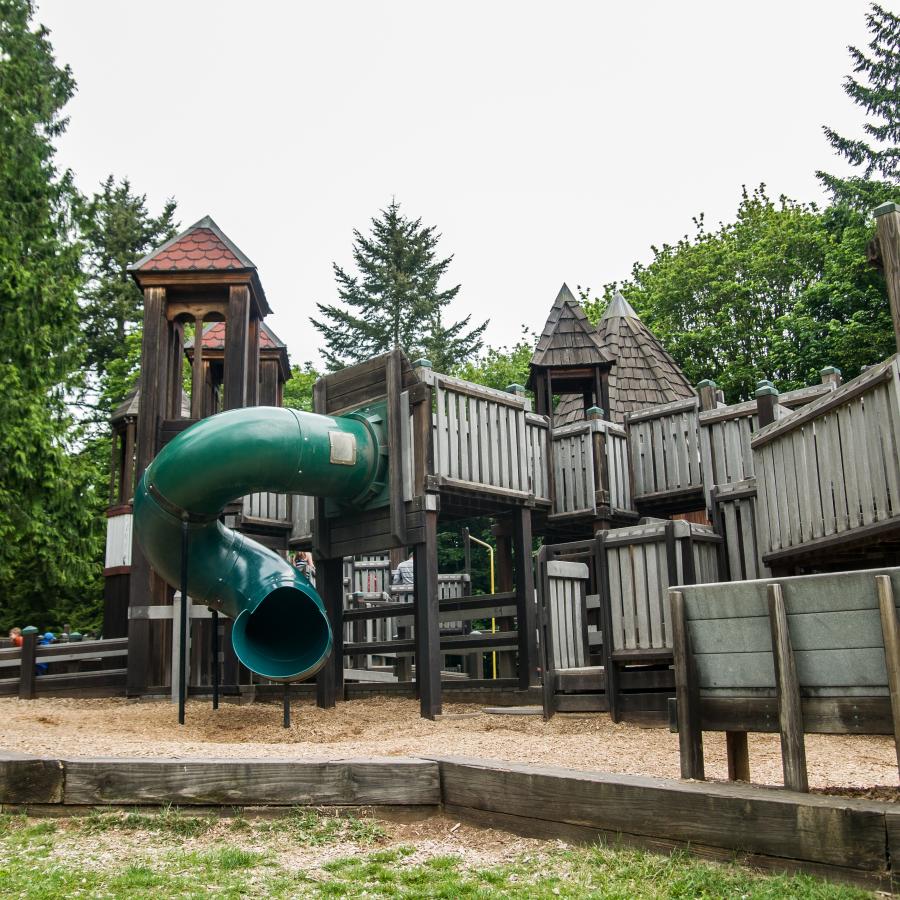 Wooden playground with a spiral slide, multiple levels of play with some pieces high in the air and others lower on the ground. 