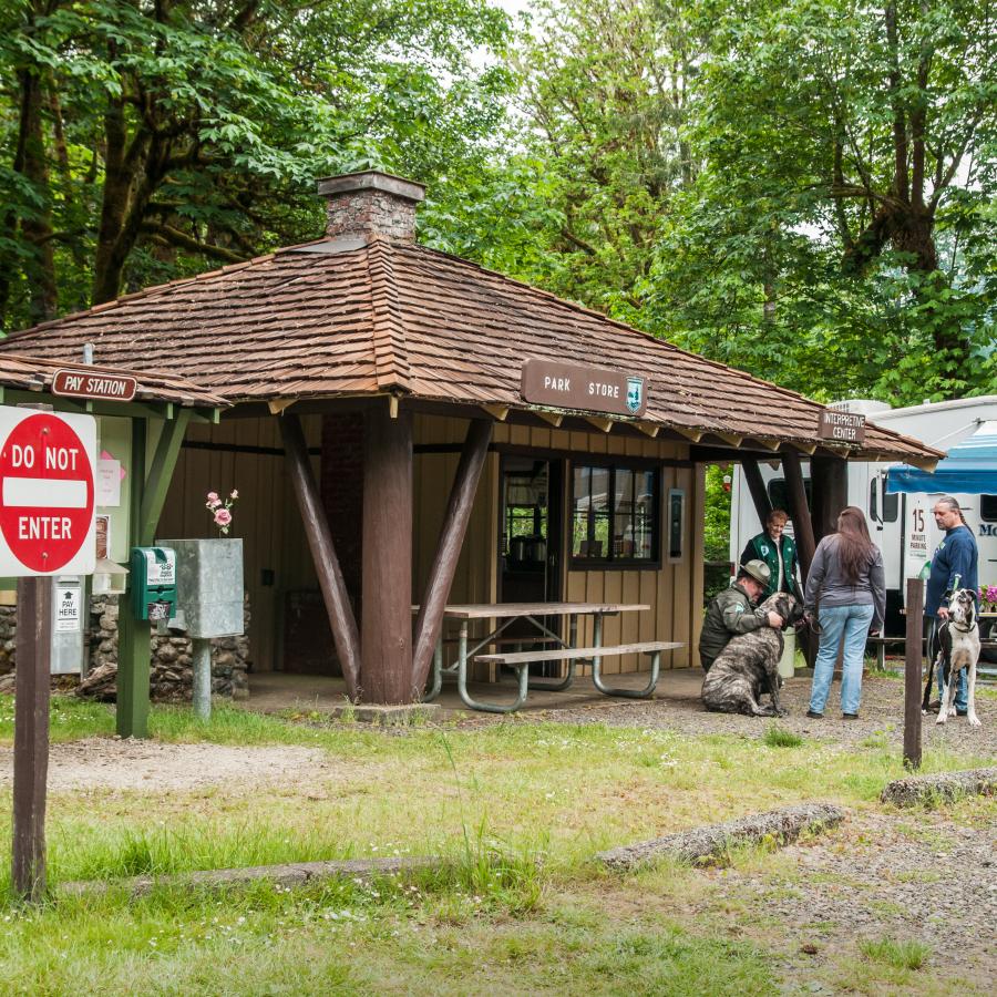 Visitors, a parks employee, and a volunteer are interacting outside the park store. This small store with yellowish-brown wooden siding and a wood shingled roof. There is a green kiosk to the left of the park store. To the right is a recreational vehicle that appears to be camping.