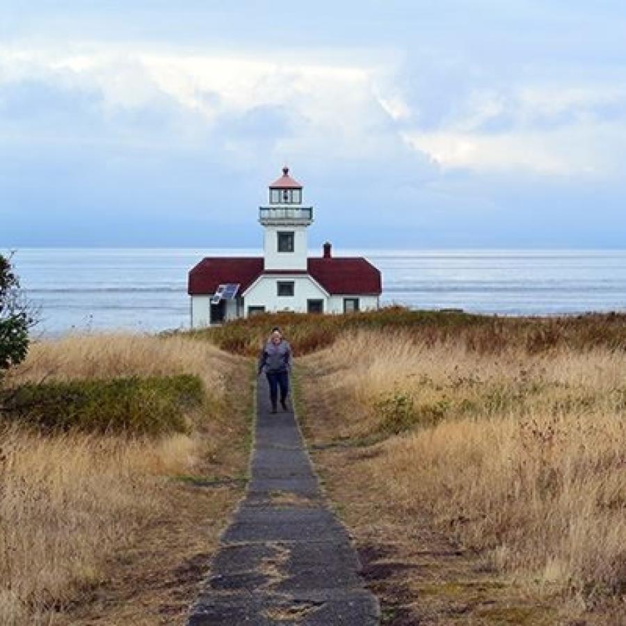 Lighthouse with white siding and a red roof centered in the photo with a trail leading up to the entrance. The ground cover is dormant, likely in early spring, and the sky is a brilliant baby blue with fluffy white clouds.