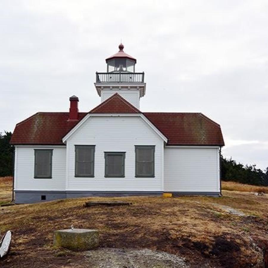 White Lighthouse with red roofing. There are 4 windows on the side facing the camera. There is one rectangular ground floor with a small second floor where the lighthouse light is. There is a railing around the light portion and a pointed red roof.
