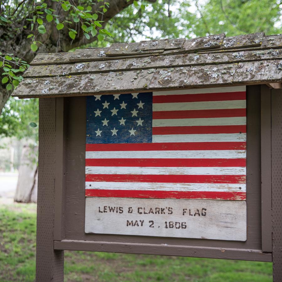 Brown roofed sign with a painted replica of Lewis and Clark's flag from 1806 with 15 stars on a blue background and 15 red and white stripes