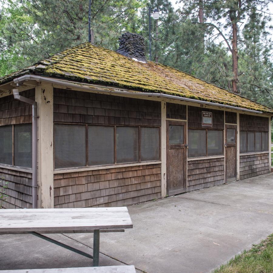 Picnic shelter with screened windows and wood shake shingles on all sides, a concrete path surrounds the shelter with three picnic tables