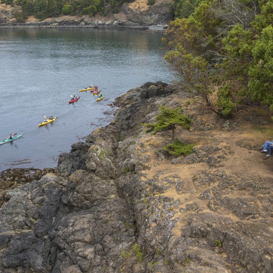 A group of six kayaks paddle along the shoreline of the rocky cliff as two hikers take a rest at a picnic table to watch the kayakers pass by. There are green trees and shrubs around the onlookers.