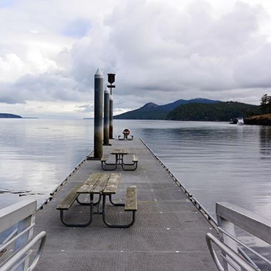 Standing on a dock with picnic tables lined down the dock. Grey waters and sky surround the dock, treed hillsides are seen in the background.
