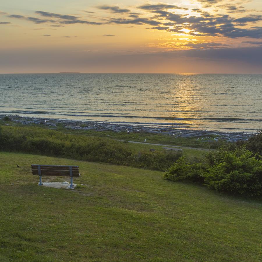 A lone bench sits in a grassy lawn looking over the water as the sun sets, creating a blue, orange and pink sky with some clouds covering the sun.