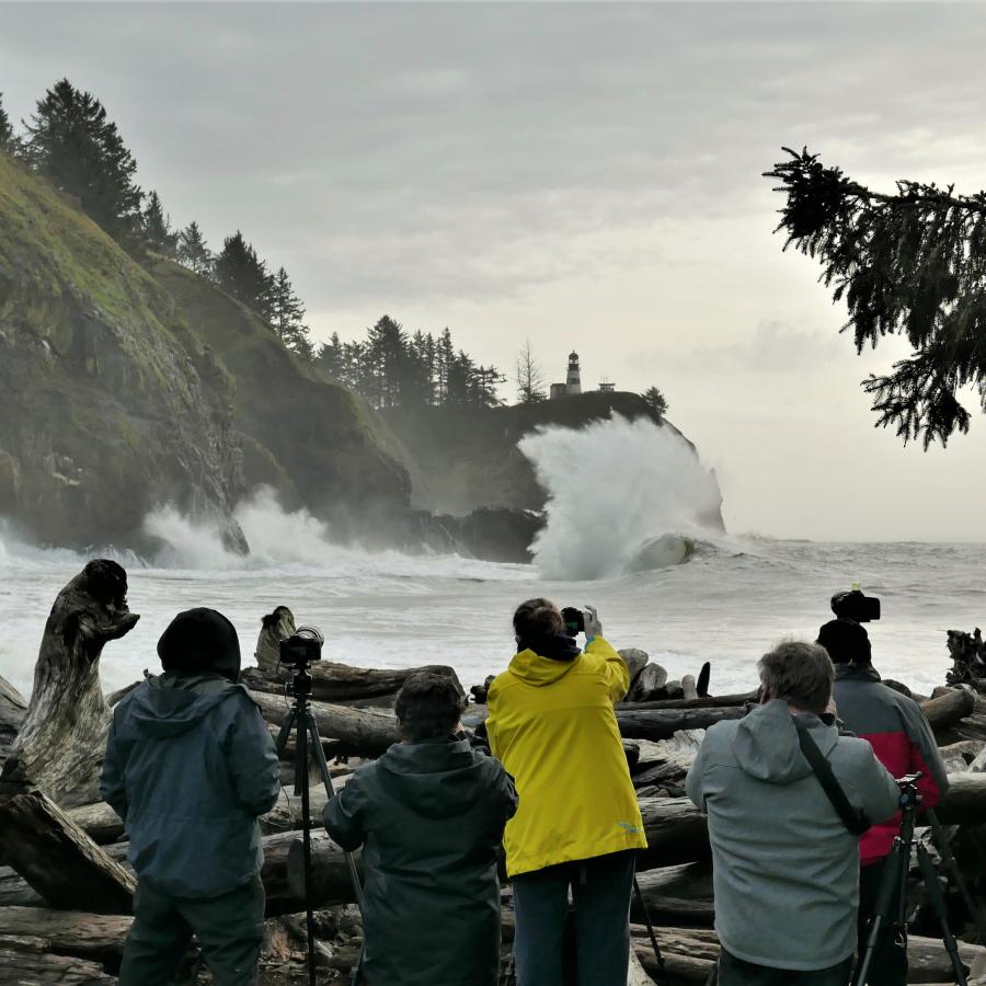 Eight people stand on a rugged shoreline holding cameras and photographing the surf crashing high against the cliffs at Cape Disappointment State Park. North Head Lighthouse can be seen in the distance on top of the cliff where the tall white wave is crashing hardest.