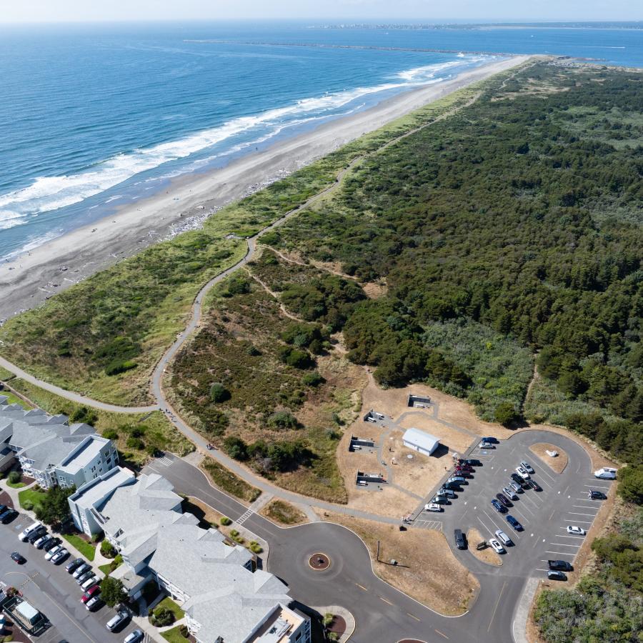 Overhead view of Westport Light State Park with beach and ocean, a winding concrete trail through grassy dunes with beachfront condos, and a forested parking area.