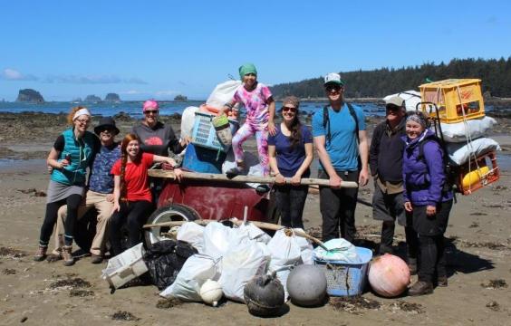 Nine volunteers standing on the beach around garbage in bags, broken buoys, a tire and more that was collected. A blue sky, ocean and small islands are seen in the background.