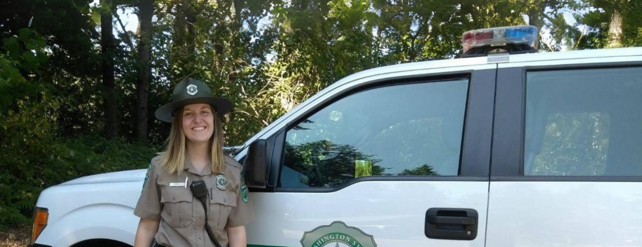 Woman stands in front of a truck with evergreen trees in the background
