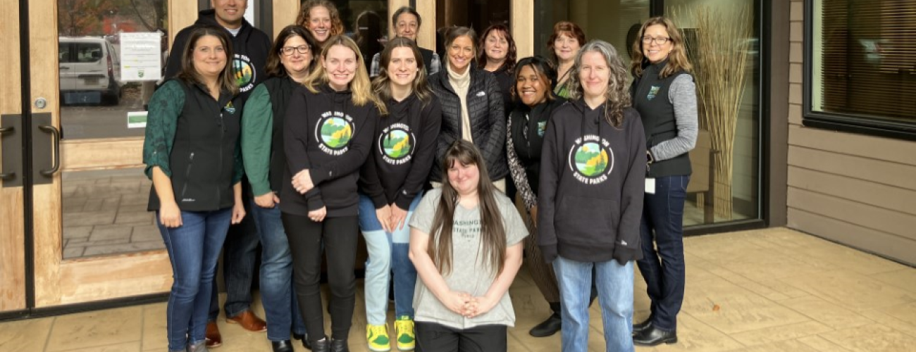 A group of people who work for the Washington State Parks human relations department posing in front of wood and glass entrance doors to the Park's headquarters building.