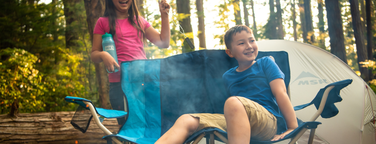 Boy sitting in a campchair with a girl behind him holding a roasting stick with a tent in the background