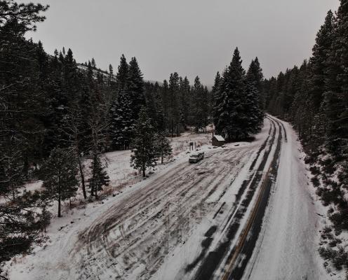 A plowed parking pull-off area next to a plowed highway flanked by snow-dusted pine trees.