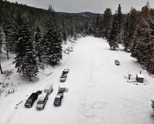 Several trucks parked on a long snow-covered parking area surrouned by pine trees. A pit toilet hut sits in the lower right corner.