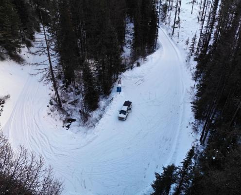 A forest road covered in thick snow and flanked by evergreen trees curves around a bend. A porta-potty and truck stand on the side of the road.