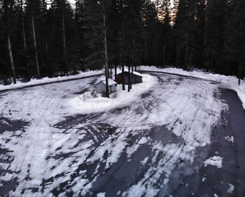 Oval paved parking lot covered thinly with snow and ice bordered by piled up snow and thick pine trees. A warming hut and pit toilet hut stand in the middle.