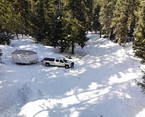A snowy forest road with a pull-out surrounded by pine trees. A truck with a trailer stands in the pull-out.
