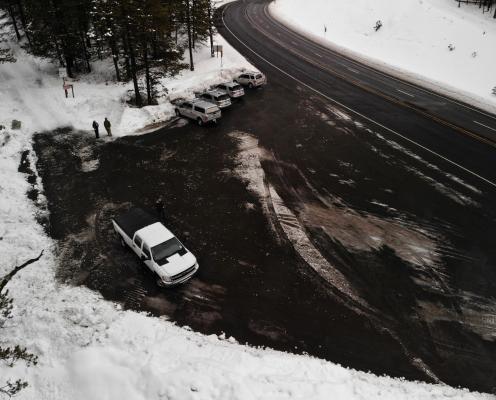 Parking area next to a highway, both plowed clean. A snowy forest lies to the left of the parking area.