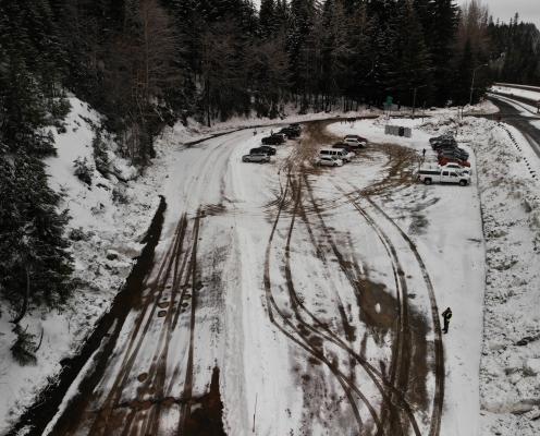 Cars parked in a parking area covered by a thin layer of snow next to a plowed highway. Five porta-potties lie just inside the parking lot entrance.