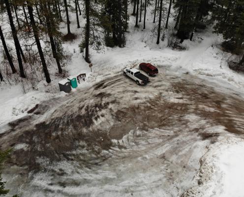 Overhead view of a plowed highway flanked by tall pine trees and snow-covered ground leading to a small plowed pull-off parking area.