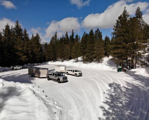 Two trucks with trailers sit in the middle of a plowed parking area covered by a layer of snow. A porta-potty stands on the right edge.