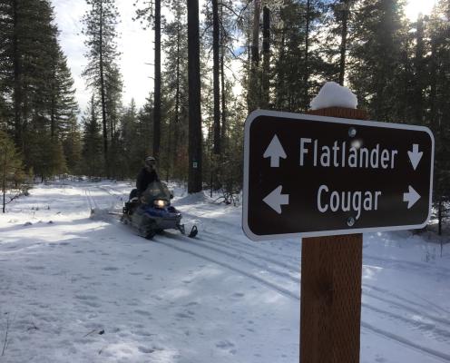 A person riding a snowmobile down a snow-covered path. A sign for Flatlander and Cougar trails is in the foreground.