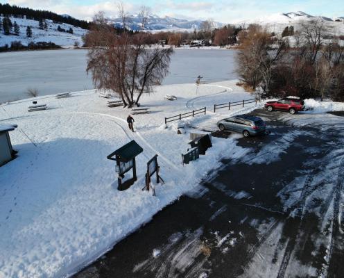 Plowed parking lot next to a snow-covered picnic area, overlooking a frozen lake with snow-covered mountains in the background.