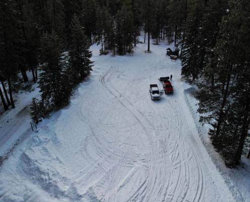 Overhead view of a snow-covered parking area lined with pine trees next to a plowed highway. A pit toilet hut sits in the upper right corner.