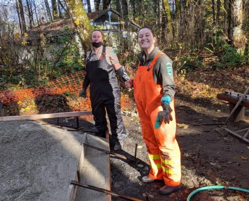 Two park aides, one in an orange waterproof jumpsuit, work on a dirt trail project with shovels and hoses.