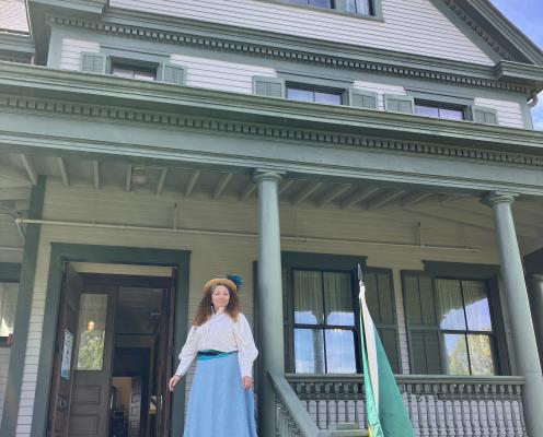 A woman in a period costume stands in front of a historic building.