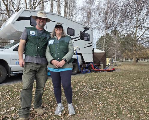 A couple stand in front of an RV in a field with trees.