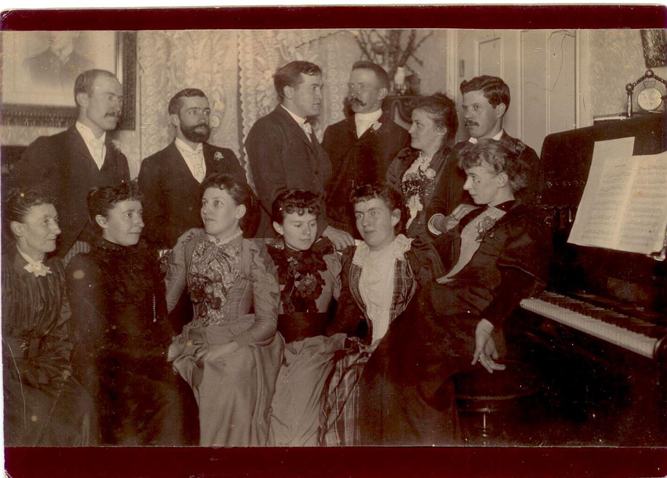 An old photo of husbands and wives in formal clothes sitting near a piano during a party.