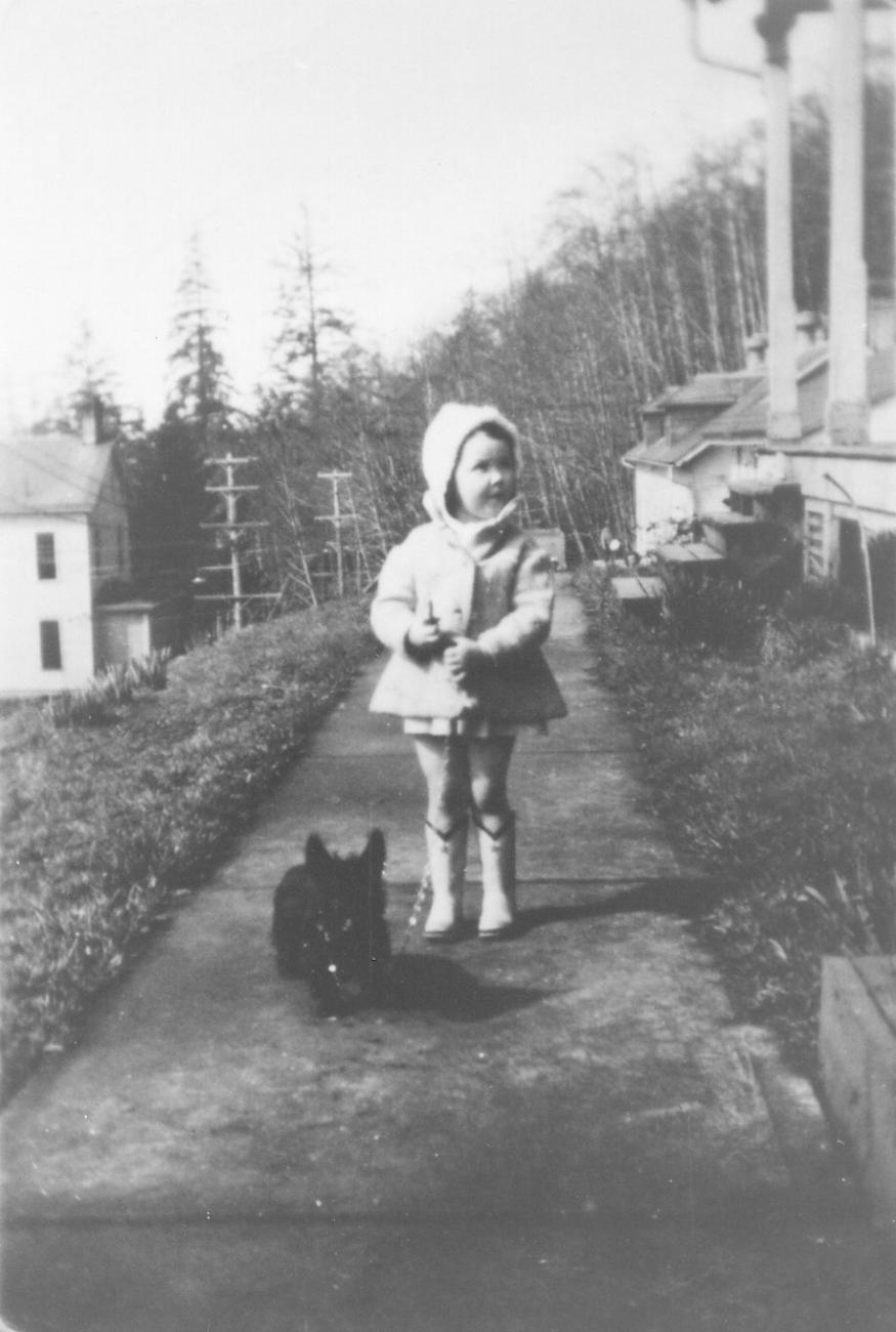 An old photo of a little girl and her little dog on a sidewalk near military housing.