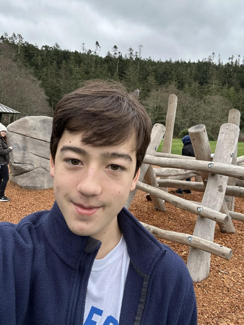 A teenage boy smiles in front of a play structure that was designed by his Youth Leadership Program at Deception Pass State Park. The structure is made of criss-crossing smooth wooden logs over wood chips with a climbing boulder. Trees surround the area.