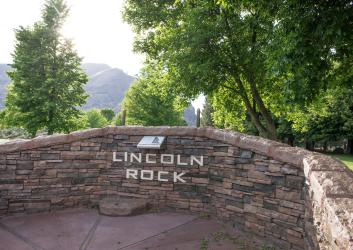 Rock wall structure with 'Lincoln Rock' with tall, leafy trees surrounding the structure. In the distance, a hillside can be seen.