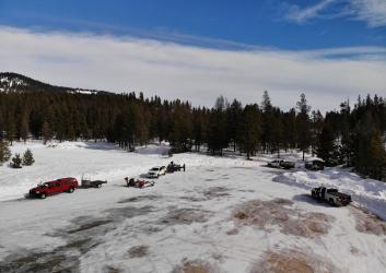 Large plowed parking lot with vehicles and snowmobiles and a pit toilet hut in the corner.