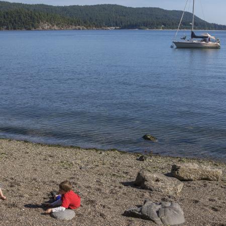 Image of two youths playing on the rocky beach in the foreground. In the midground a sailboat can be seen on the water. The boat is white with a red stripe horizontally across the body. The sails are down. In the background, land across the water is visible with forested hills.