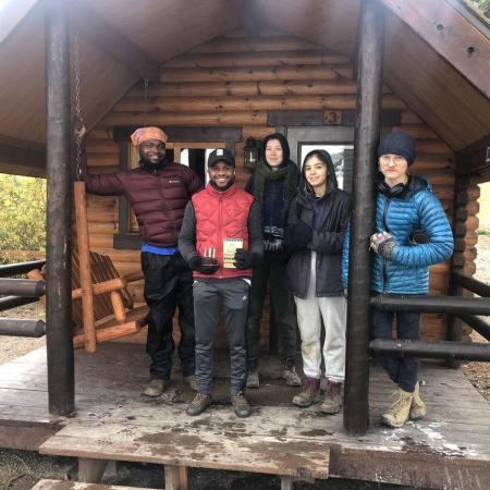 Five people celebrate a birthday in font of a wood cabin with a dusting of snow