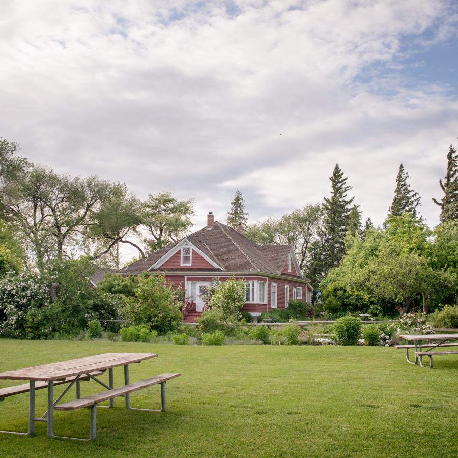 Red house built in 1908 hidden in the middle of pine trees, lilac and rose bushes. Manicured lawn with picnic tables. Large white and grey clouds in the sky with blue sky coming through.