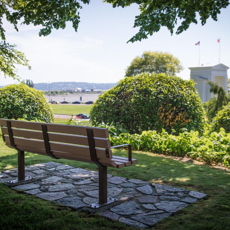 Sitting bench in the shade on a sunny day looking out at the manicured flower beds and ocean in the distance.