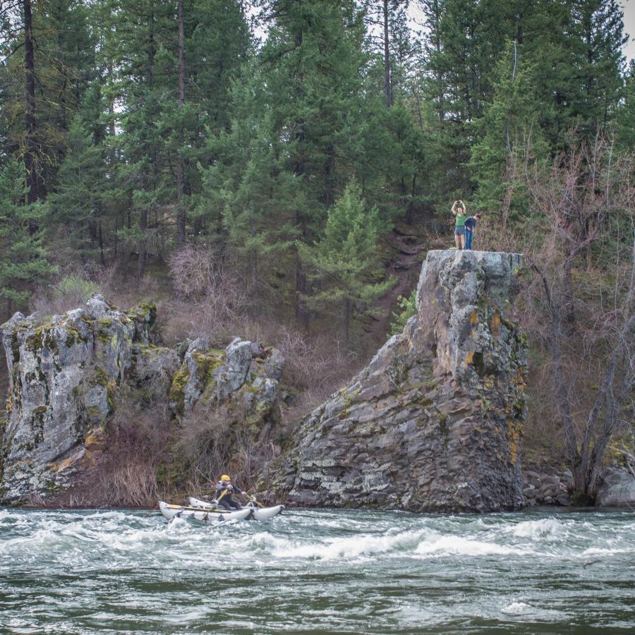 A boat rows through a river rapid while two visitors look on atop a cliff on the far side of the river.
