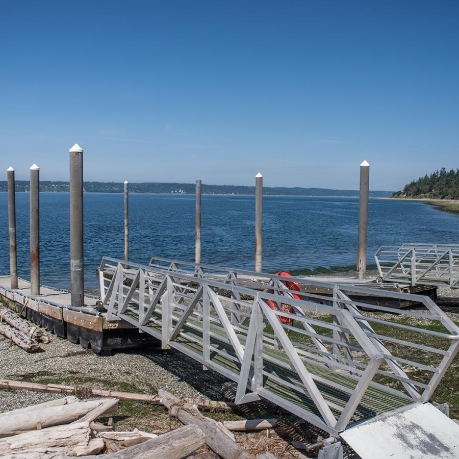 Boat Launch at Camano Island with two metal gangways. There is driftwood on the rocky beach and trees on a rocky incline along the beach on the right side.