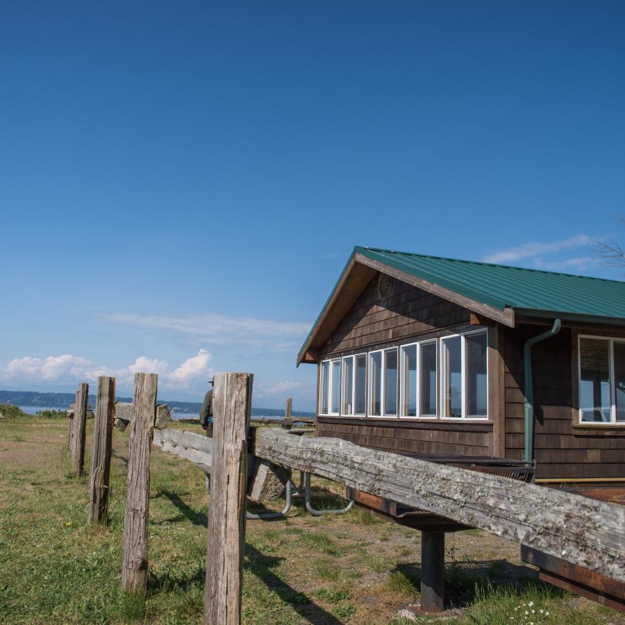 Picnic shelter at Camano Island State Park. The shelter has a green, metal-looking roof, dark brown wooden siding, 5 white windows along the left side that take up the entire left wall, a single window visible on the entrance side of the building and a large, white framed door visible. The building is next to a picnic table, grill, and visible wooden post fence. In the far background you can just make out the Saratoga Pass and land on the opposite side of the water.