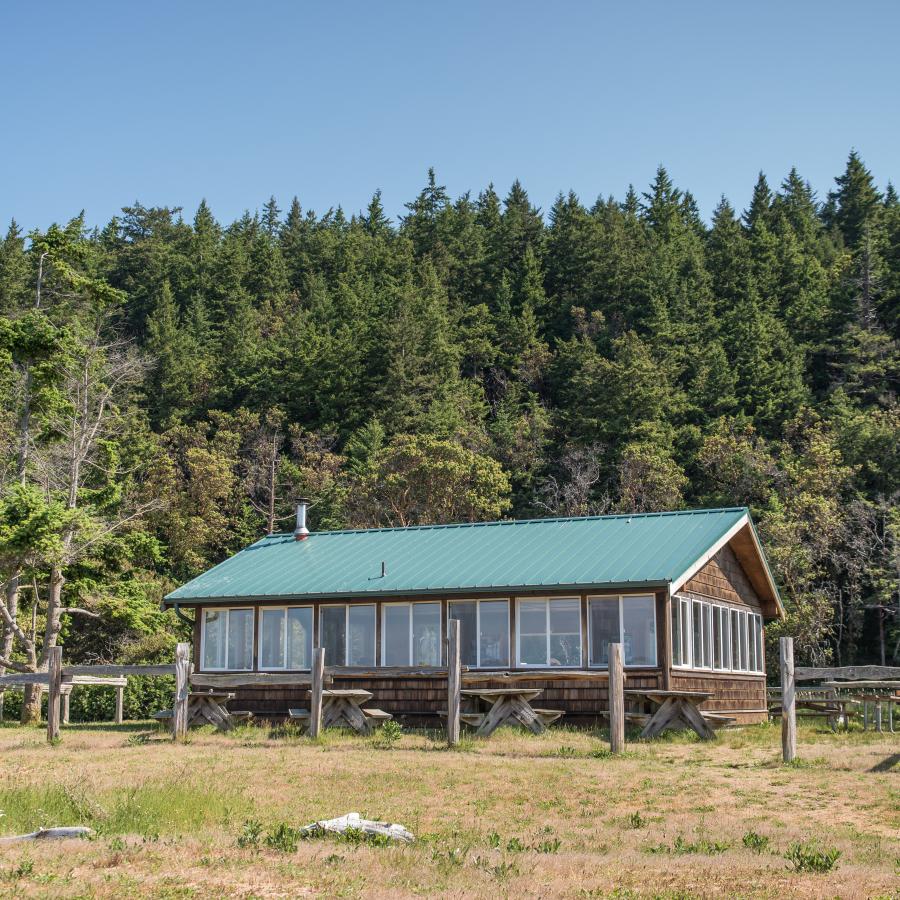 Back and left side of the picnic shelter exterior. The back of the picnic shelter has 7, large, white windows that fill the entire back side. Along the left side of the building there is 5 white-framed windows which span the entire left side of the building. The roof is metal-looking and green and the siding is a dark brown stained wood. There is a wood post-fence visible with some of the fencing materials missing so that all you see is posts. There are picnic tables visible along both sides.