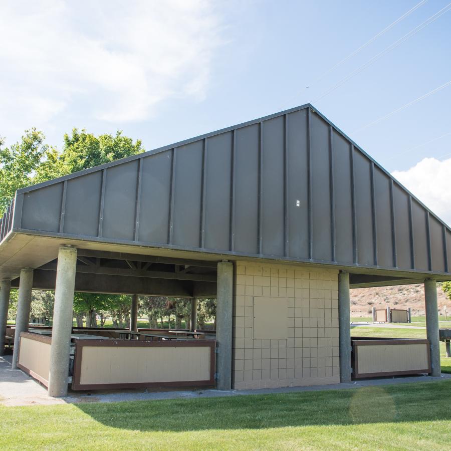 A large picnic shelter sits on a lawn with rows of trees behind it.