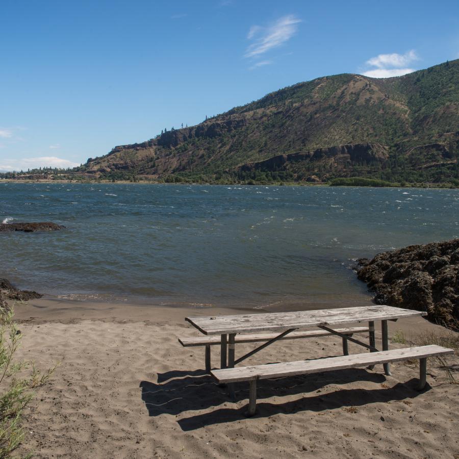 Sandy beach with picnic table next to shoreline lake cliffs in the distance and green boat guide marker light